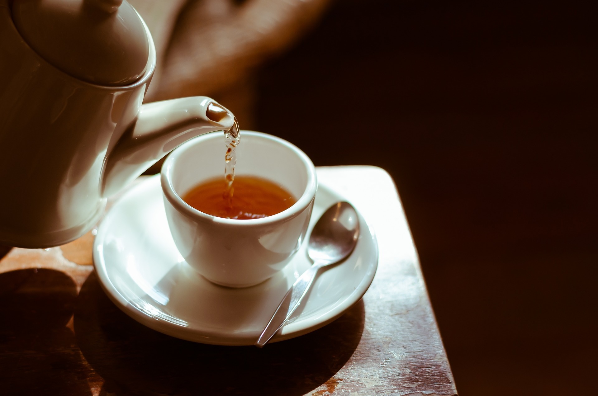 Tea being poured from a white teapot into a cup on a sunlit wooden table