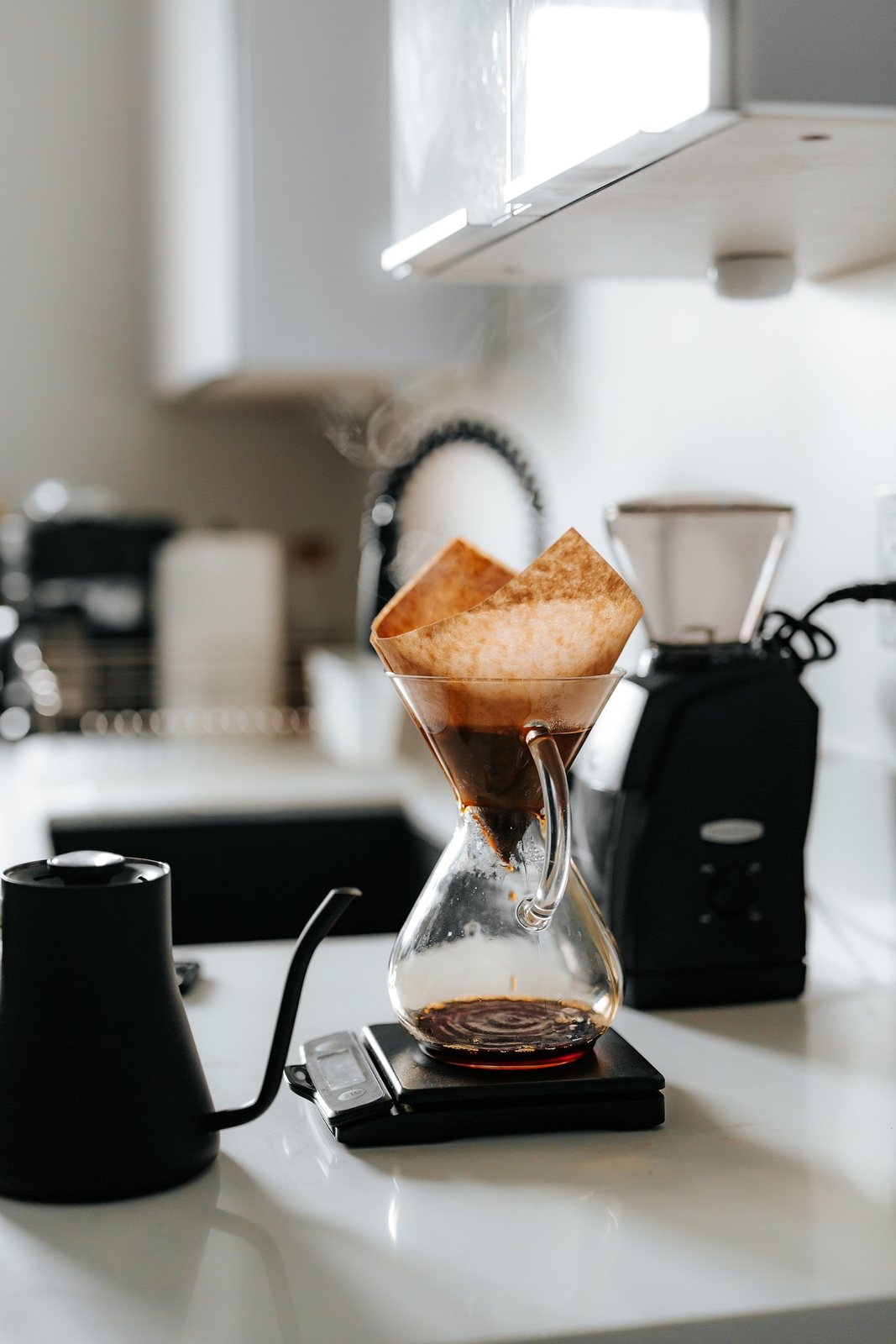 Coffee brewing through a brown paper filter into a glass carafe beside a kettle and grinder